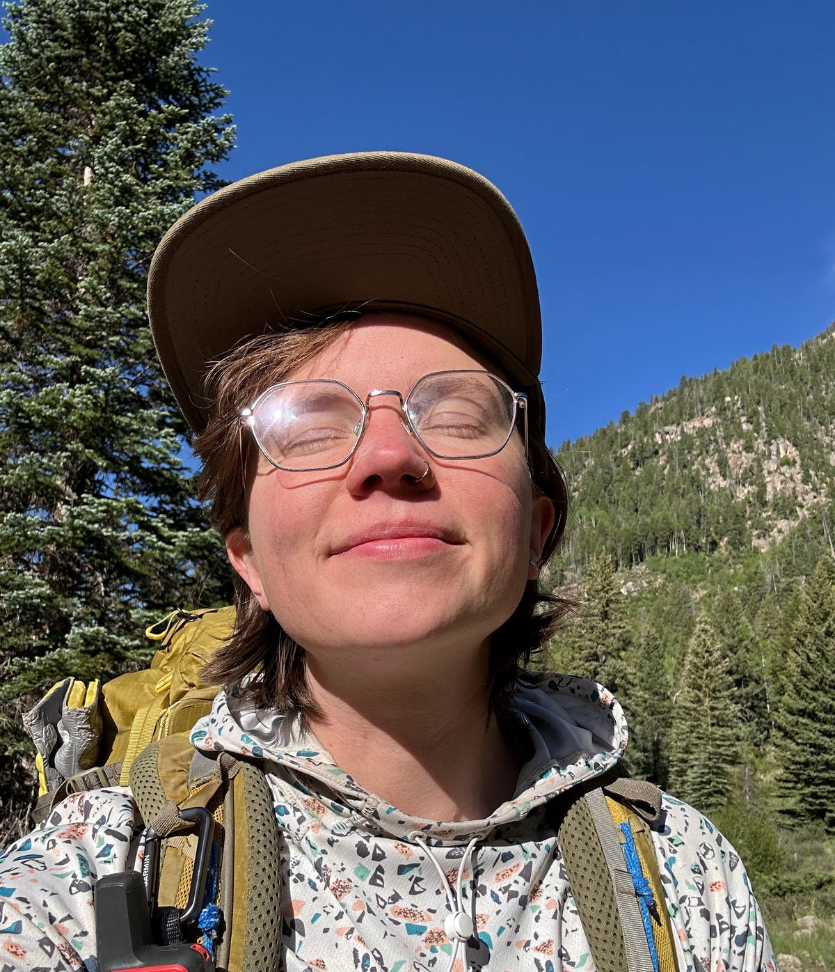 Person with glasses and a cap standing outdoors with mountains and trees in the background